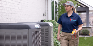 A high-resolution photograph of a female 911 Mechanical technician in a navy blue polo and khaki cargo pants, walking past residential AC condenser units while holding a diagnostic tool. Her white cap and shirt chest display the 911 Mechanical logo. The brick wall of a house and garden are in the background.