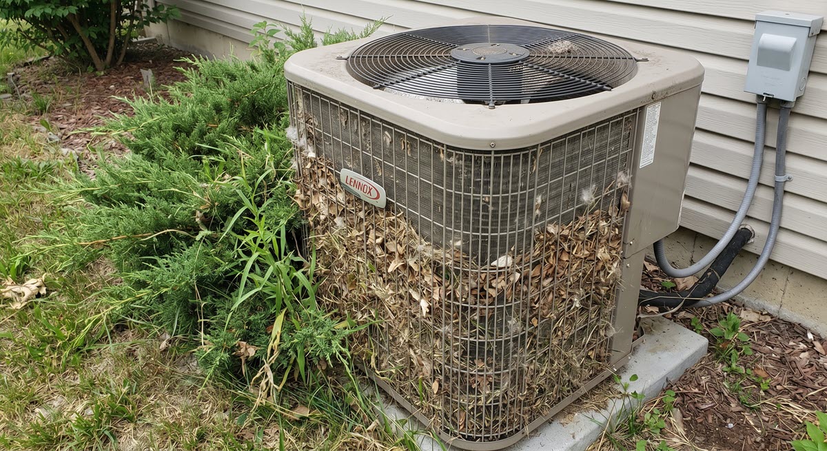 An outdoor Lennox air conditioning unit heavily clogged with dead leaves and debris, with a green bush growing closely against its left side.